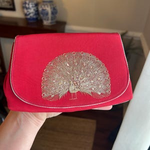 K&ouml;nnte beinhalten: Eine rote Clutch mit einem goldenen gestickten Pfauenmuster.