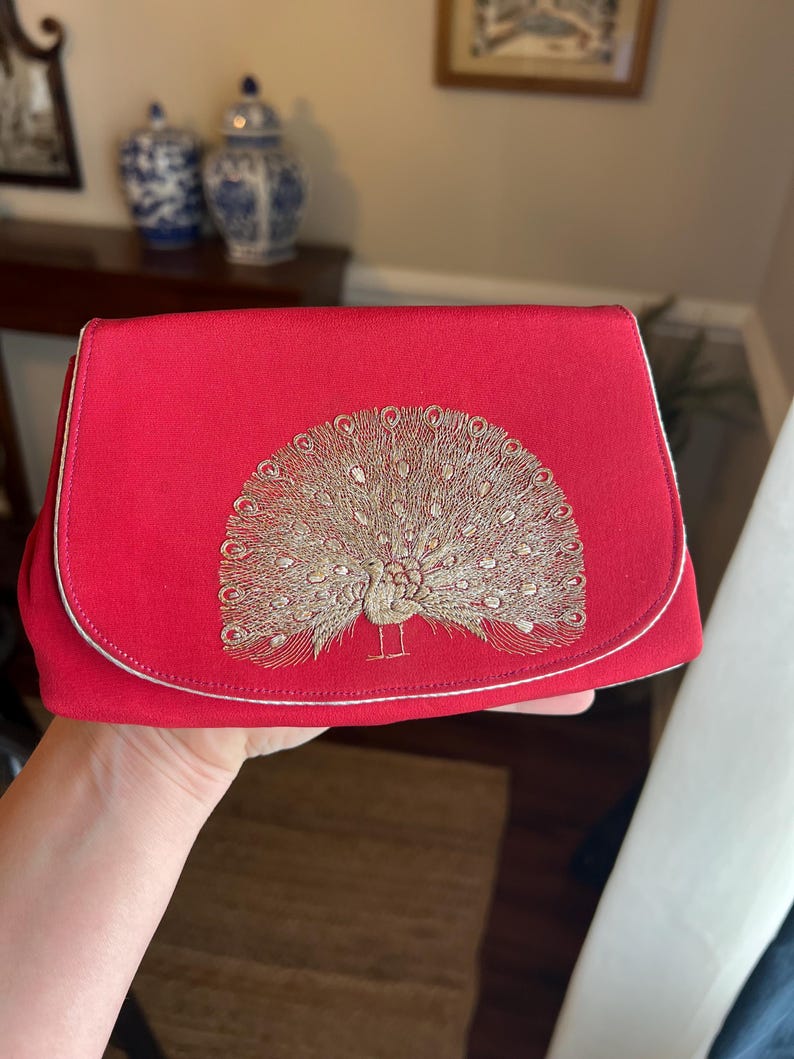 K&ouml;nnte beinhalten: Eine rote Clutch mit einem goldenen gestickten Pfauenmuster.
