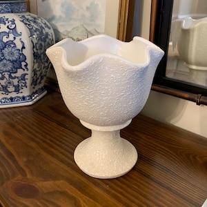 May include: White ceramic vase with a textured surface and a scalloped rim. The vase has a pedestal base.