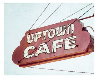 May include: A vintage red and white neon sign with the words "Uptown Cafe" hanging from chains against a light blue sky.