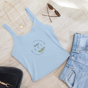 Pode incluir: Um top sem mangas azul claro com um círculo branco e o texto "choose peace" em letras pretas. Uma samambaia verde está dentro do círculo. O top está sobre uma superfície de madeira clara com um par de óculos de sol e uma calça jeans azul.