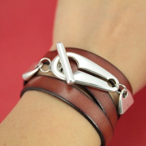 May include: A brown leather wrap bracelet with a silver toggle clasp. The bracelet is made of three strands of leather and has a silver charm hanging from the clasp.