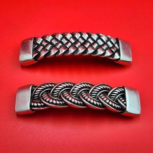 May include: Two silver-colored metal decorative pieces with braided and woven designs. The top piece has a woven pattern, while the bottom piece features a braided rope design. Both pieces have rectangular end caps against a red background.