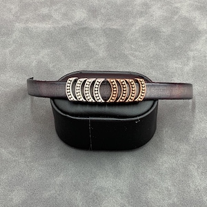 May include: A brown leather bracelet with a silver and gold metal charm featuring a series of half-circles with small beads.