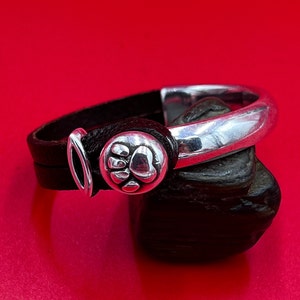 May include: A black leather bracelet with a silver paw print charm and a silver clasp. The bracelet is wrapped around a dark gray stone against a red background. The bracelet is approximately 0.5 inches wide.