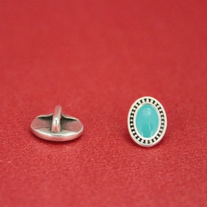 May include: A silver oval button with a turquoise blue center and a decorative border. The button has a shank on the back for sewing.