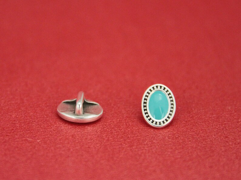 May include: A silver oval button with a turquoise blue center and a decorative border. The button has a shank on the back for sewing.