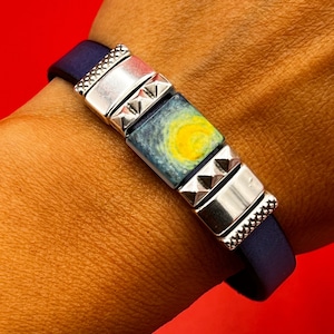 May include: A navy blue leather bracelet with silver accents and a square tile featuring a painting of a starry night sky with a yellow sun.