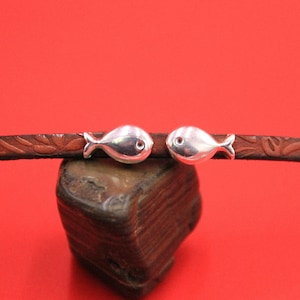 May include: A brown leather bracelet with two silver fish charms. The bracelet is on a brown rock.
