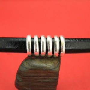 May include: A close-up of a black leather cord with a silver metal bead accent. The bead is made up of several stacked rings.