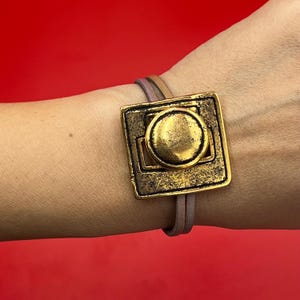 May include: A gold-toned bracelet with a square-shaped charm featuring a circle design. The bracelet is made of brown leather.