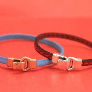 May include: Two leather bracelets, one light blue and one red, with metal clasps. The bracelets are displayed on a red background. The clasps are a contrasting color to the bracelet bands.