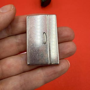 May include: A silver-toned rectangular lighter with a curved handle on the front. The lighter is held in a hand against a red background. A small, dark stone is visible in the background.