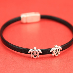 May include: A black leather bracelet with two silver turtle charms. Each turtle charm has a peace sign design.