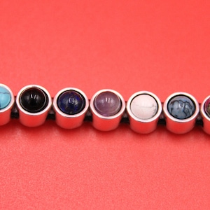 May include: A leather bracelet with eight interchangeable round metal charms. Each charm features a different colored gemstone, including turquoise, black, blue, purple, white, gray, and green.
