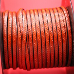 May include: A spool of brown leather cord with a decorative pattern. The cord is wound tightly around a metal spool.