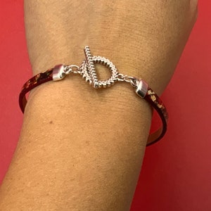 May include: A red leather bracelet with a silver toggle clasp. The bracelet has a textured pattern and is worn on a person's wrist.