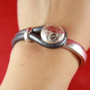 May include: A black leather bracelet with a silver clasp and a silver button with an engraved design.