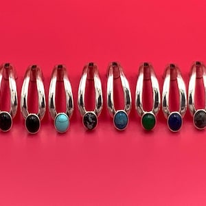 May include: Eight silver-toned metal rings arranged in a horizontal row. Each ring features a split-shank design and a single oval-shaped cabochon gemstone in various colors, including black, turquoise, blue, green, and dark grey.