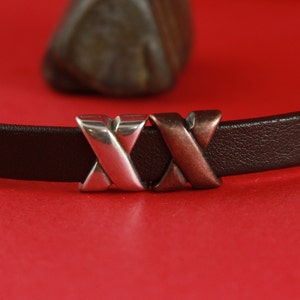 Peut inclure: Un bracelet en cuir marron avec deux breloques en forme de 'X' en argent et en cuivre.