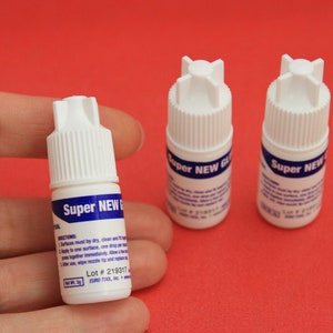 May include: Three small white bottles of Super New Glue on a red surface. The bottles have blue labels with white text. The label on the bottle in the center reads "Super New Glue" and includes instructions and a lot number.