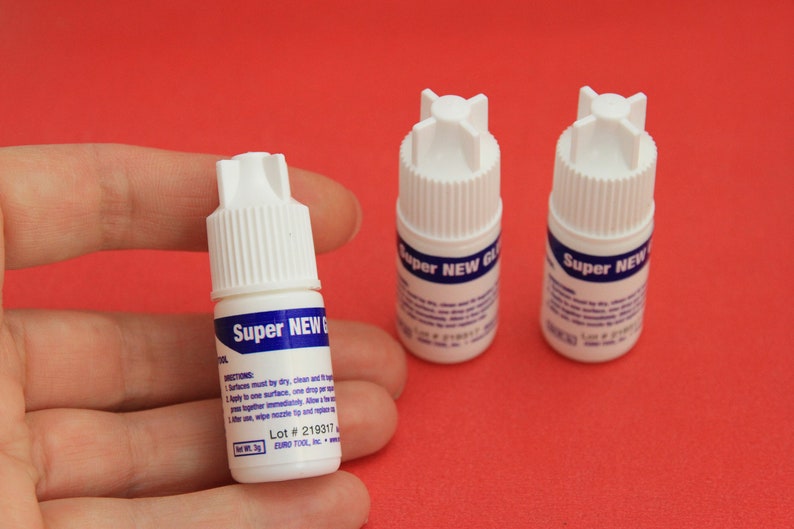 May include: Three small white bottles of Super New Glue on a red surface. The bottles have blue labels with white text. The label on the bottle in the center reads "Super New Glue" and includes instructions and a lot number.