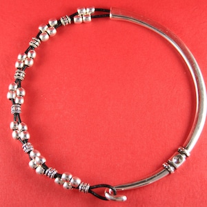 May include: Silver-toned bracelet with a curved, open design. The bracelet features a black cord adorned with silver beads and decorative elements. The clasp is a simple hook and loop closure. The jewelry is set against a red background.