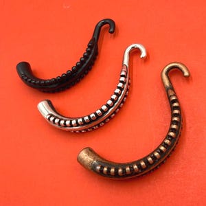 May include: Three curved, tentacle-shaped jewelry components arranged diagonally. One is solid black, one is metallic silver, and one is a metallic bronze finish. Each piece features a row of small, raised circular bumps along the inner curve.