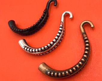 Könnte beinhalten: Drei gebogene, tentakelförmige Schmuckkomponenten, die diagonal angeordnet sind. Eines ist einfarbig schwarz, eines ist metallisch silber und eines hat ein metallisches Bronze-Finish. Jedes Teil hat eine Reihe kleiner, erhabener runder Noppen.