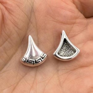 May include: Two silver-toned, fan-shaped pendants. One pendant features a Greek key design along the bottom edge. The other pendant has a textured interior. Both have a loop at the top for attaching to a chain or other jewelry.