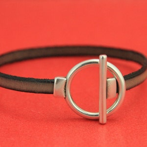 May include: A black leather bracelet with a silver toggle clasp. The clasp is a circle with a bar across the center.