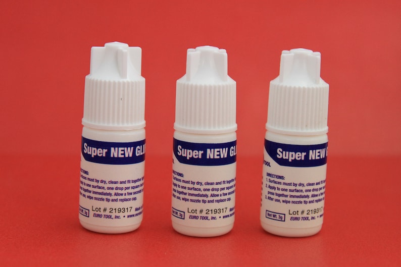 May include: Three white bottles of Super New Glue with blue labels and white caps. The bottles are on a red surface. The labels have instructions for use and a lot number: Lot # 219317.