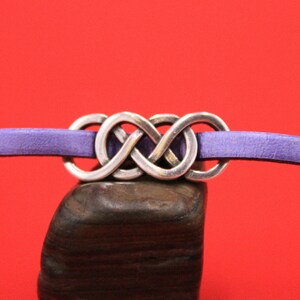 May include: A silver infinity knot bracelet with a purple leather strap. The infinity knot is the central feature, set against a red backdrop. The bracelet is displayed on a dark, textured surface.