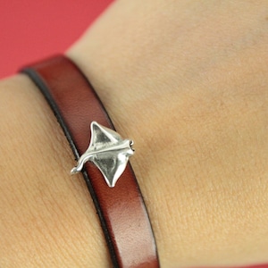 May include: A brown leather bracelet with a silver stingray charm. The bracelet is a thin band of dark brown leather. The stingray charm is a detailed, three-dimensional design. The bracelet is worn on a wrist.