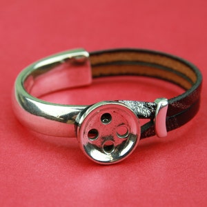 May include: A silver bracelet with a large, silver button charm. The bracelet is made of two black leather straps.