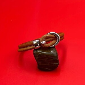 May include: A brown leather bracelet with a silver-colored clasp and a large silver ring. The bracelet is resting on a dark, speckled stone against a bright red background. The bracelet appears to be approximately 8 inches in circumference.