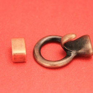 May include: A close-up of a metal clasp with a loop and a hook. The clasp is a dark bronze color and is on a red surface. There is a small, square, copper-colored piece to the left of the clasp.