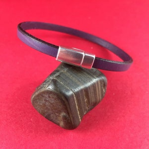 May include: A purple leather bracelet with a silver magnetic clasp. The bracelet is displayed on a dark, textured stone against a red background. The bracelet is a simple, elegant design.