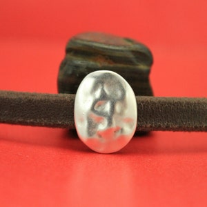 May include: A silver oval-shaped bead with a hammered texture. The bead is on a brown leather strap.