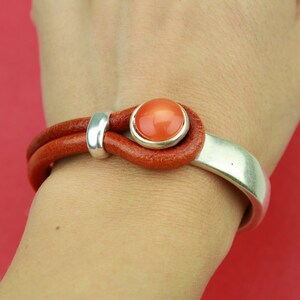 May include: A brown leather bracelet with a silver metal band and a round orange gemstone. The bracelet is worn on a person's wrist.