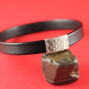 May include: A black leather bracelet with a silver clasp featuring a geometric pattern.