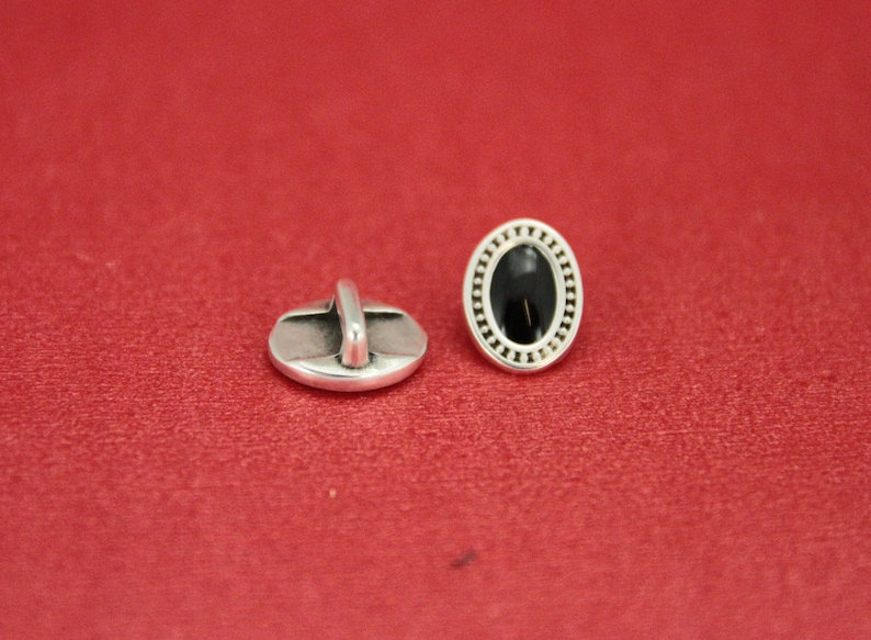 May include: Two silver oval buttons with a black center. One button has a decorative border and the other has a loop on the back.