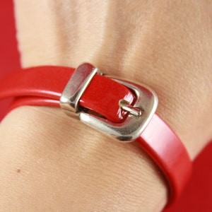 May include: A red leather bracelet with a silver buckle. The bracelet is wrapped around a wrist.