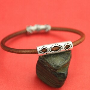 May include: A brown leather bracelet with a silver charm featuring a geometric design.