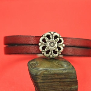 May include: A double-layered leather bracelet with a silver-toned floral charm. The bracelet is red and the charm has a detailed design.