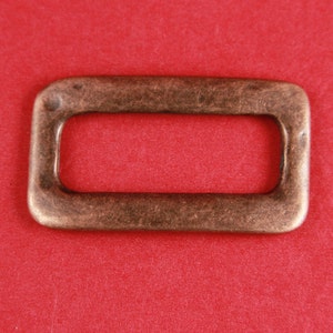 May include: A rectangular metal buckle with a slightly rounded edge, finished in a copper tone.