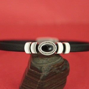 May include: A black leather bracelet with a silver oval charm featuring a black gemstone. The charm is surrounded by a decorative border of small silver beads.
