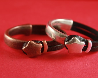 Könnte beinhalten: Zwei Lederarmbänder mit metallischen Stern-Akzenten. Ein Armband ist silberfarben mit schwarzem Leder und das andere ist kupferfarben mit schwarzem Leder.