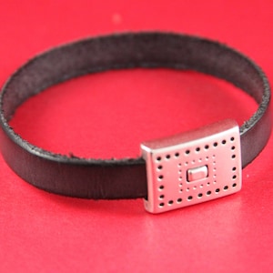May include: A black leather bracelet with a silver clasp featuring a square design with a smaller square in the center.