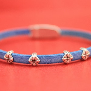 May include: A blue leather bracelet with four silver-colored decorative accents. The bracelet has a silver clasp and is set against a red background. The accents have a cross-like design.